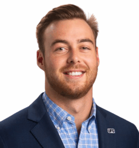 A male business consultant wearing a blue blazer, checkered shirt, and smiling, with short brown hair.